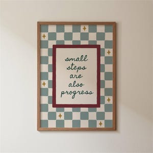 May include: A framed print with the words "small steps are also progress" in green script. The print has a checkered background in light blue and cream, with gold star accents. The frame is a light brown wood.