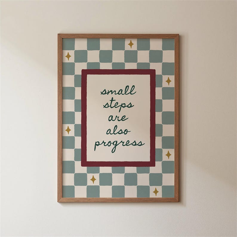 May include: A framed print with the words "small steps are also progress" in green script. The print has a checkered background in light blue and cream, with gold star accents. The frame is a light brown wood.