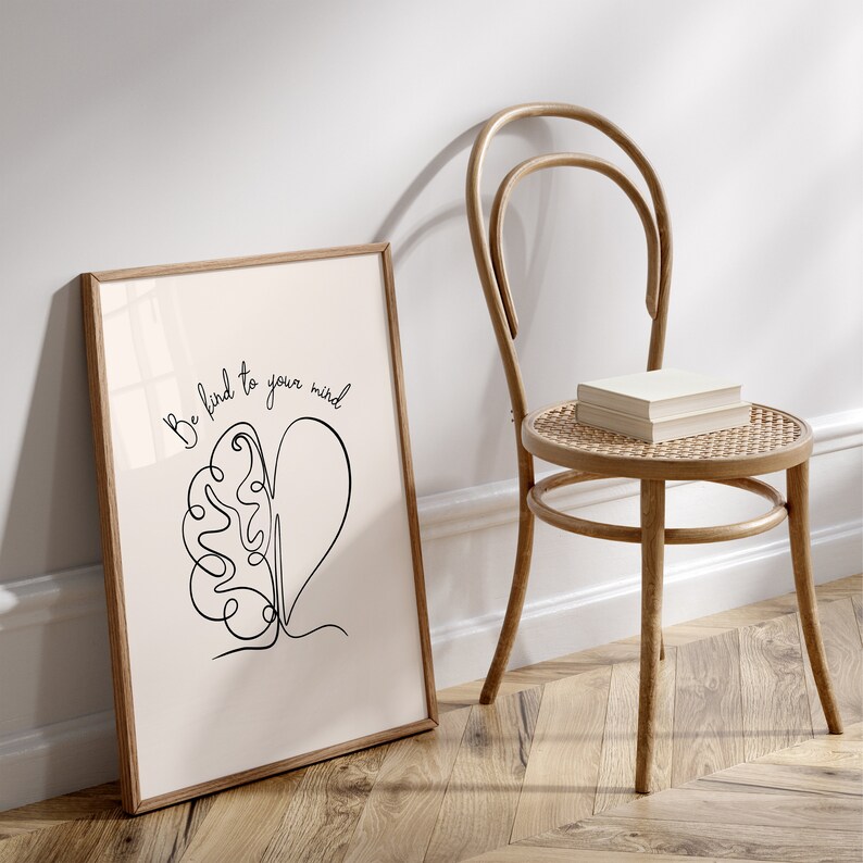 May include: A framed print with a white background and black line art of a brain and heart with the text "Be kind to your mind". The print is leaning against a wooden chair with a woven seat.