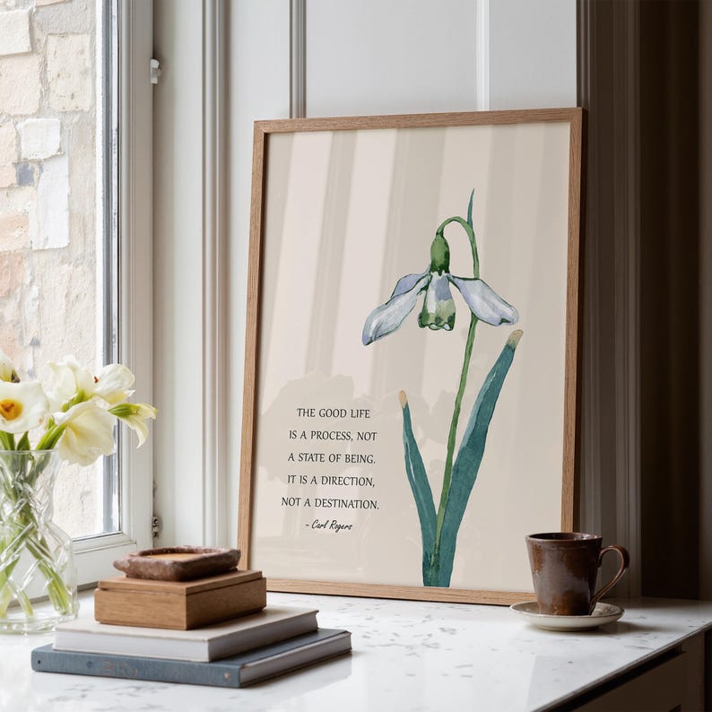 K&ouml;nnte beinhalten: Ein gerahmter Kunstdruck mit einer Aquarellillustration einer Schneegl&ouml;ckchenblume, gr&uuml;nen Bl&auml;ttern und einem hellbeigen Hintergrund. Der Druck enth&auml;lt das Zitat "The Good Life is a process, not a state of being..." von Carl Rogers.