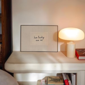 May include: A framed print with the text "how lucky are we" sits on a white shelf, next to a cream-colored mushroom-shaped lamp. A red book is also on the shelf. The print has a black frame and a dotted border around the text.