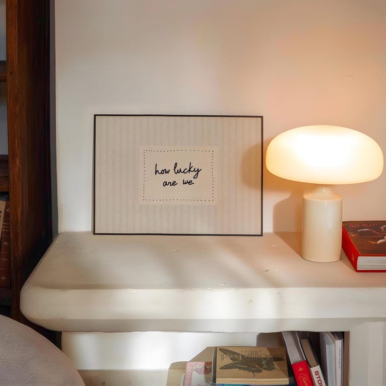 May include: A framed print with the text "how lucky are we" sits on a white shelf, next to a cream-colored mushroom-shaped lamp. A red book is also on the shelf. The print has a black frame and a dotted border around the text.