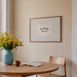 May include: A framed artwork with the text "how lucky are we" hangs on a beige wall. A round wooden table holds an open book, a brown mug, and a light blue vase with yellow flowers. A wooden chair is positioned near the table.