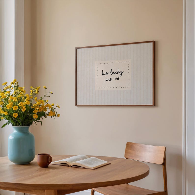 May include: A framed artwork with the text "how lucky are we" hangs on a beige wall. A round wooden table holds an open book, a brown mug, and a light blue vase with yellow flowers. A wooden chair is positioned near the table.