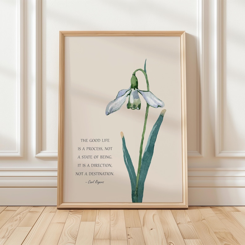 K&ouml;nnte beinhalten: Gerahmter Kunstdruck mit einer Aquarellillustration einer Schneegl&ouml;ckchenblume in Blau- und Gr&uuml;nt&ouml;nen. Der Druck enth&auml;lt das Zitat "The Good Life is a Process, Not a State of Being" von Carl Rogers. Der Rahmen ist hellbraun.