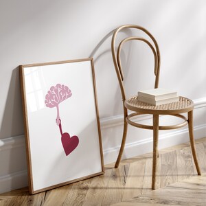 Puede incluir: Una impresi&oacute;n enmarcada con una silueta rosa de un &aacute;rbol cuyas ramas alcanzan una mano que sostiene un coraz&oacute;n. La impresi&oacute;n est&aacute; sobre un suelo de madera junto a una silla de madera.