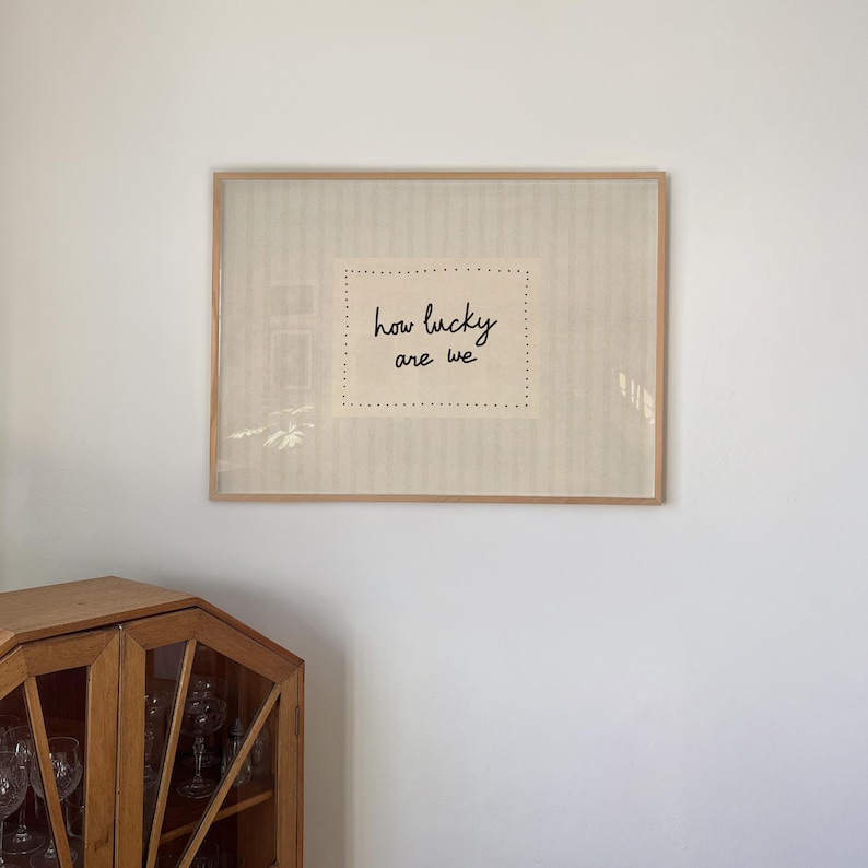 May include: A framed artwork with the text "how lucky are we" in black script. The artwork is set against a beige background with a dotted border, encased in a light brown wooden frame, and mounted on a white wall.
