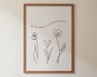 May include: A framed wall art print featuring a white background with black line art illustrations of three distinct flowers. Above the flowers, the text "WE ALL GROW AT DIFFERENT RATES" is printed in a simple, black, sans-serif font.