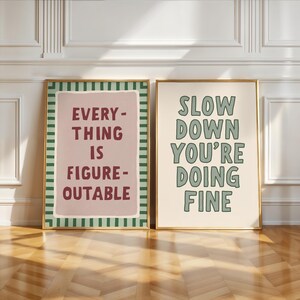 May include: Two framed art prints with inspirational quotes. One print reads "EVERYTHING IS FIGURE-OUTABLE" in burgundy text on a pink background. The other says "SLOW DOWN YOU'RE DOING FINE" in green on a cream background.