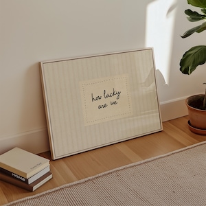 May include: A framed artwork with the text "how lucky are we" in a handwritten style. The artwork is in a neutral color palette and leans against a white wall. A stack of books and a potted plant are also visible.