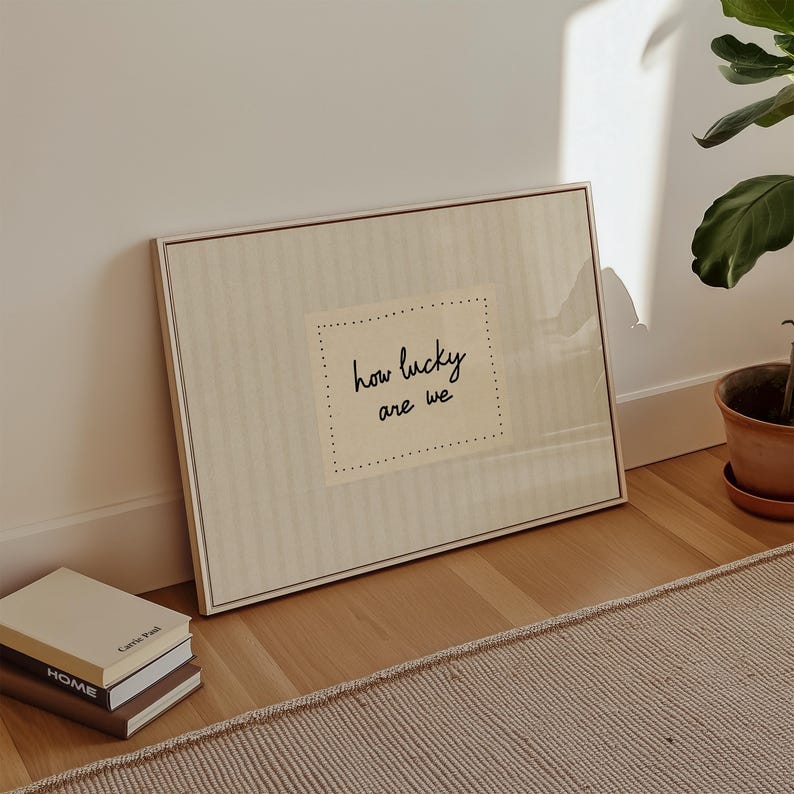 May include: A framed artwork with the text "how lucky are we" in a handwritten style. The artwork is in a neutral color palette and leans against a white wall. A stack of books and a potted plant are also visible.