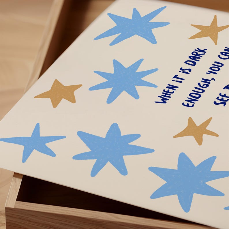 May include: A cream-colored print featuring blue and gold stars. The text "WHEN IT IS DARK ENOUGH, YOU CAN SEE THE STARS" is written in navy blue. The print is displayed in a wooden drawer.