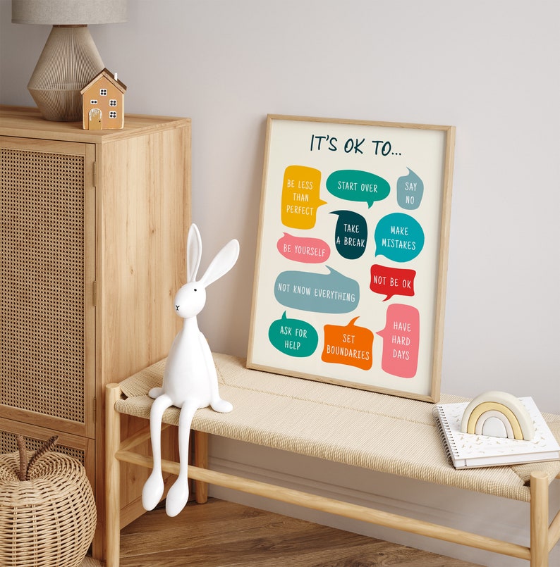 May include: A framed print with the text "IT'S OK TO..." and various phrases in colourful speech bubbles. The print is displayed on a light-coloured bench, alongside a white rabbit figurine and a wooden cabinet.