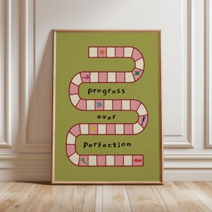 Op de afbeelding: Een ingelijste wandkunstprint met een olijfgroene achtergrond en een kronkelend, roze en wit bordspelpad. De tekst "progress over perfection" is in het zwart gedrukt. Het pad bevat kleine iconen zoals een ster, vraagteken en een hartje.