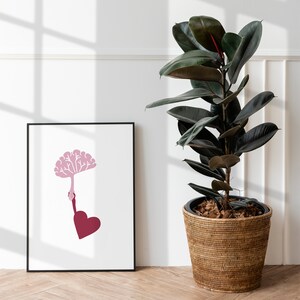 Puede incluir: Una impresi&oacute;n enmarcada en negro con un &aacute;rbol estilizado de color rosa que crece de una mano y un coraz&oacute;n rojo. La impresi&oacute;n est&aacute; colgada en una pared blanca junto a una planta de caucho en maceta.