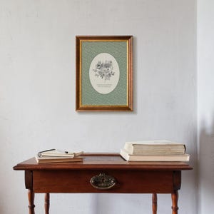 May include: A framed floral print with a gold-coloured frame hangs on a white wall. The print features a green background with a floral pattern and an oval shape with a floral illustration and text that reads "Grow through what you go through."