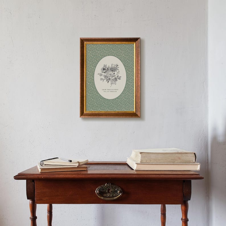 May include: A framed floral print with a gold-coloured frame hangs on a white wall. The print features a green background with a floral pattern and an oval shape with a floral illustration and text that reads "Grow through what you go through."