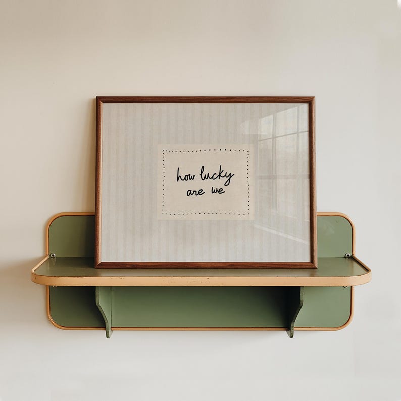 May include: A framed print with the text "how lucky are we" displayed on a green and tan shelf. The print has a brown frame and a neutral-toned background. The shelf is mounted on a white wall.