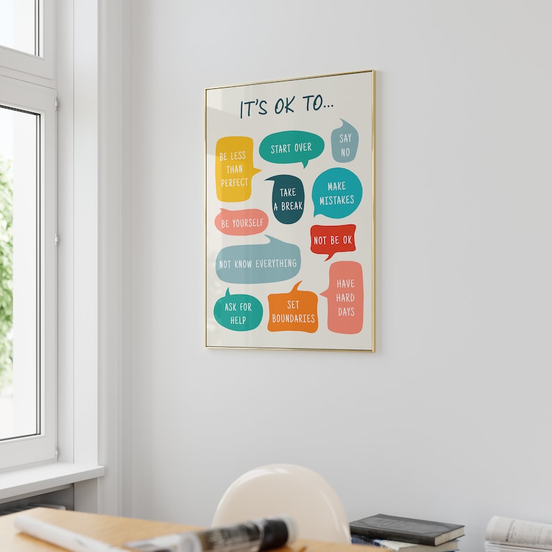 May include: A framed poster with the text "IT'S OK TO..." and various phrases in colourful speech bubbles, including "Be less than perfect," "Start over," and "Say no." The poster is in a gold frame and hangs on a white wall.