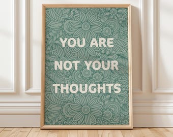 May include: A framed art print with the words "YOU ARE NOT YOUR THOUGHTS" in white lettering. The background is a teal floral pattern. The frame is a light wood color. The print is leaning against a white wall and wooden floor.