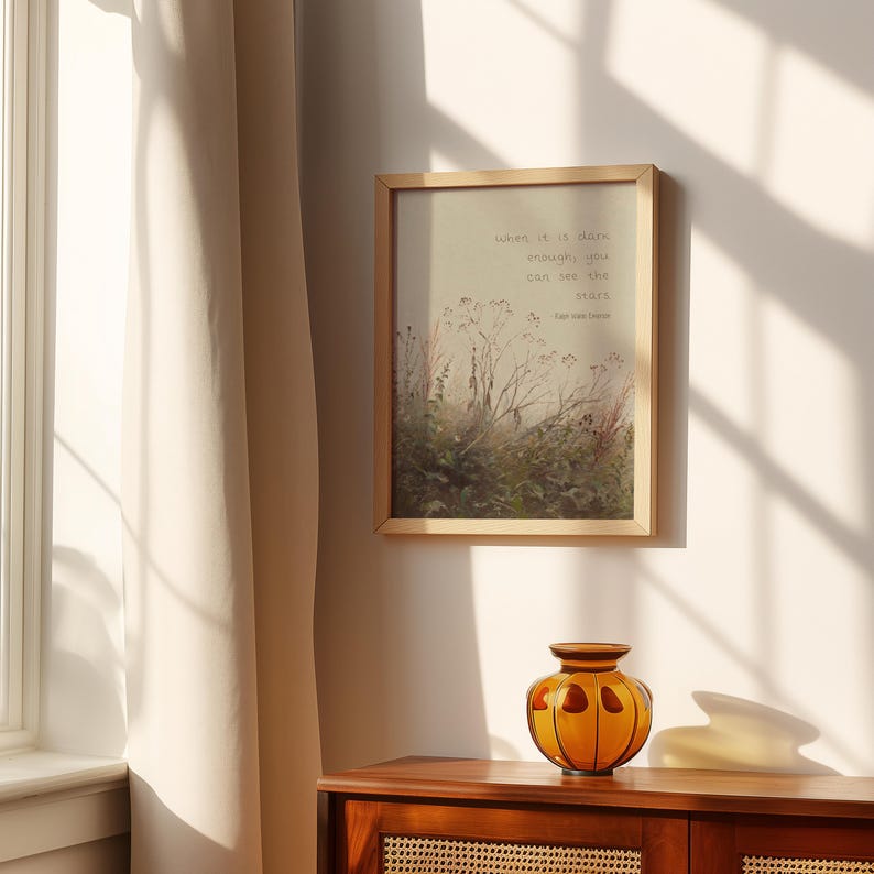 May include: A framed print with a light wood frame displays a watercolour-style landscape with text that reads, "when it is dark enough, you can see the stars." An orange glass vase sits on a wooden cabinet.