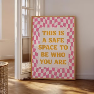 May include: A framed poster with a pink and white checkered border and the words "THIS IS A SAFE SPACE TO BE WHO YOU ARE" in yellow. The poster is displayed in a room with a wooden floor and a white door.