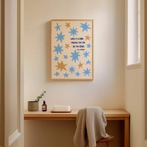 May include: A framed art print featuring blue and gold stars on a cream background. The print includes the quote "WHEN IT IS DARK ENOUGH, YOU CAN SEE THE STARS" by R.W. Emerson. The artwork is displayed above a wooden desk.