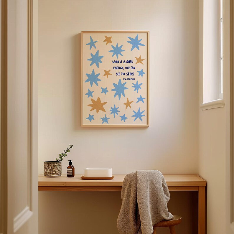 May include: A framed art print featuring blue and gold stars on a cream background. The print includes the quote "WHEN IT IS DARK ENOUGH, YOU CAN SEE THE STARS" by R.W. Emerson. The artwork is displayed above a wooden desk.