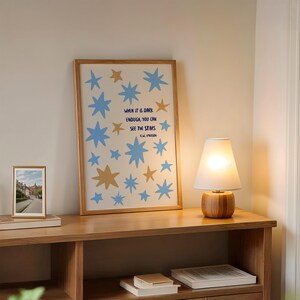 May include: A framed art print with blue and gold star illustrations and the quote, "WHEN IT IS DARK ENOUGH, YOU CAN SEE THE STARS." A small framed photo and a wooden lamp with a white lampshade sit on a wooden shelf.