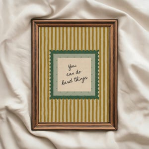 Peut inclure: Une impression encadrée avec un cadre en bois marron. L'œuvre présente un fond crème avec des rayures verticales dans des tons or et crème. Un motif carré à bordure verte au centre contient le texte manuscrit : « You can do hard things ».
