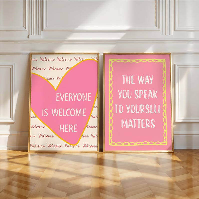May include: Two framed art prints with gold frames. One print has a pink heart with the words "EVERYONE IS WELCOME HERE." The other print has the words "THE WAY YOU SPEAK TO YOURSELF MATTERS" on a pink background.