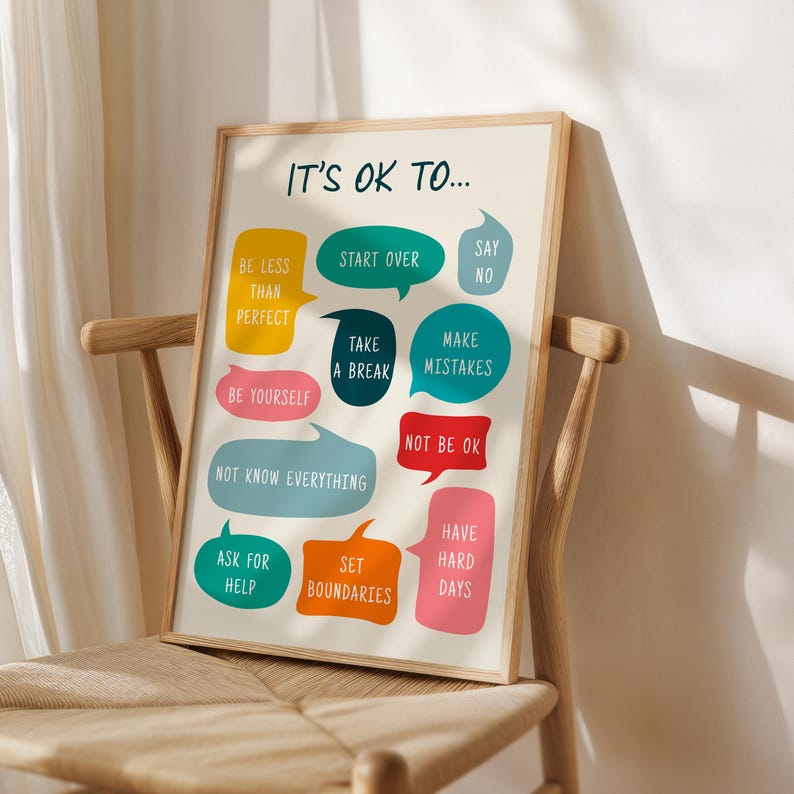 May include: A framed print with the text "IT'S OK TO..." and various phrases in colourful speech bubbles, including "Be Less Than Perfect" and "Make Mistakes." The print is displayed on a wooden chair.