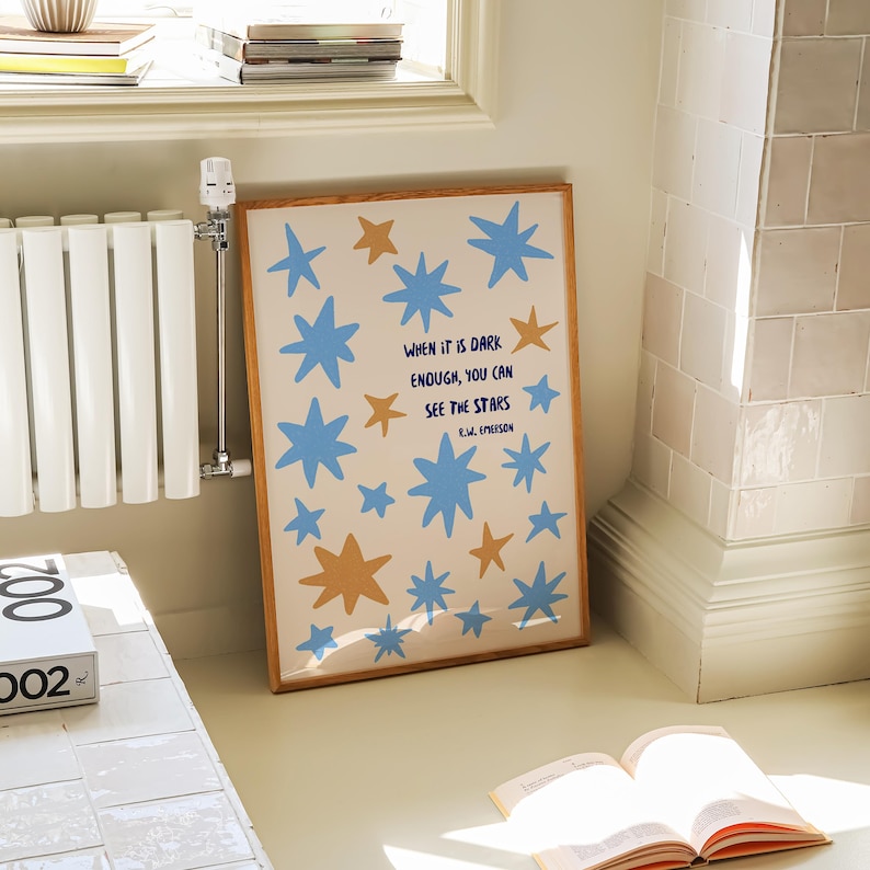 May include: A framed art print featuring blue and gold star illustrations on a cream background. The print includes the quote, "When it is dark enough, you can see the stars." The artwork is displayed on a light-colored floor.