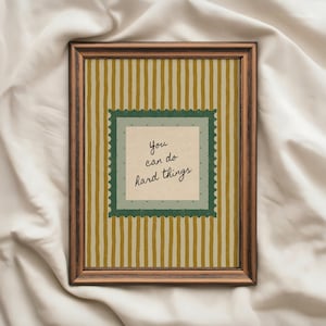 May include: A framed print with a brown wooden frame. The artwork features a cream-colored background with vertical stripes in shades of gold and cream. A green square with a scalloped edge contains the text "You can do hard things."