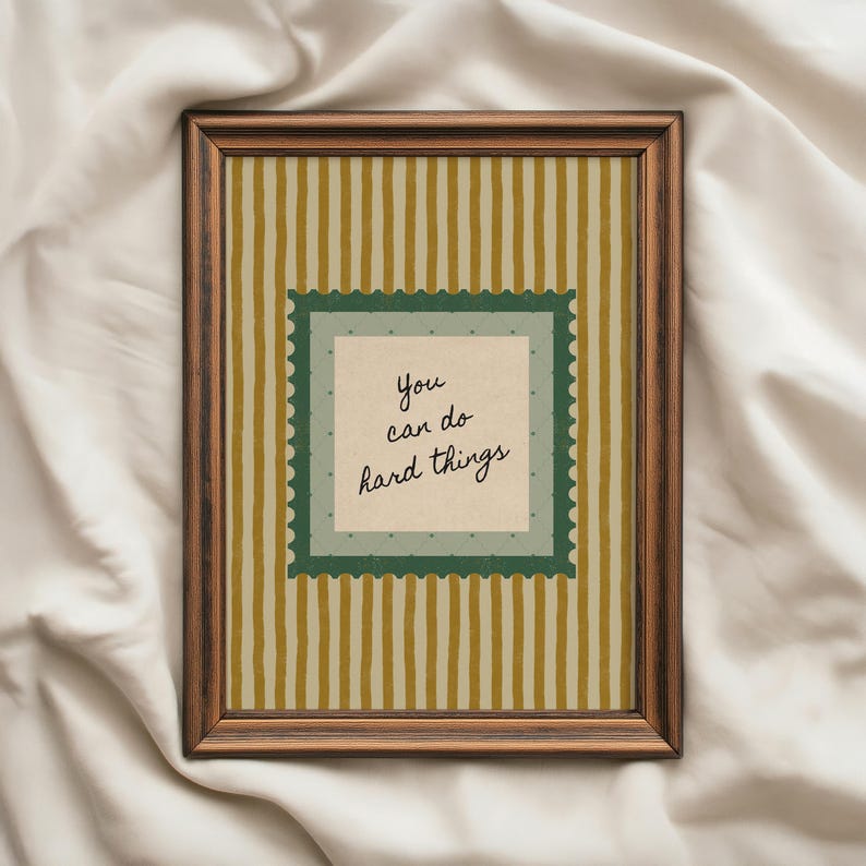 May include: A framed print with a brown wooden frame. The artwork features a cream-colored background with vertical stripes in shades of gold and cream. A green square with a scalloped edge contains the text "You can do hard things."