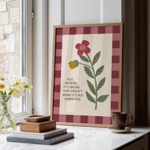 May include: A framed floral art print with a burgundy gingham border. The artwork features a stylized flower with a pink bloom and the text: "JUST BECAUSE IT'S TAKING TIME DOESN'T MEAN IT'S NOT HAPPENING". The print is displayed on a white countertop.