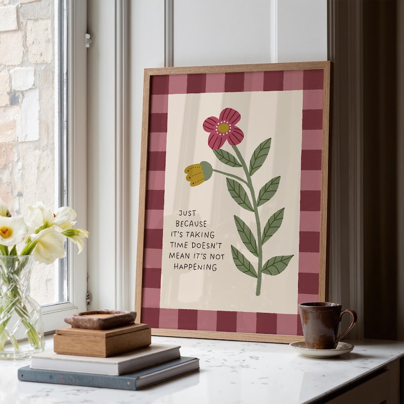 May include: A framed floral art print with a burgundy gingham border. The artwork features a stylized flower with a pink bloom and the text: "JUST BECAUSE IT'S TAKING TIME DOESN'T MEAN IT'S NOT HAPPENING". The print is displayed on a white countertop.