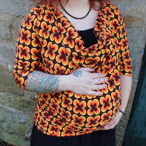 Könnte beinhalten: Oberteil mit Dreiviertelärmeln und einem Retro-Blumenmuster in Orange, Gelb und Braun. Das Oberteil hat einen Wasserfallausschnitt. Das Bild zeigt eine Nahaufnahme des Oberteils.