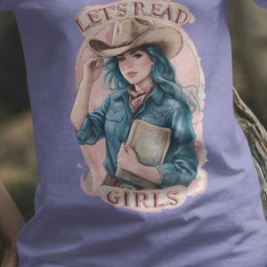 Puede incluir: Una camiseta morada con un gráfico de una mujer con un sombrero de vaquero y sosteniendo un libro. El texto "Let's Read Girls" está impreso en la camiseta.