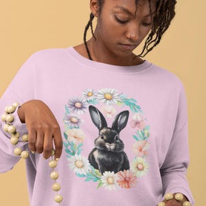 May include: A lavender colored sweatshirt with a black rabbit surrounded by a floral wreath. The wreath is made up of pink, white, and purple flowers.