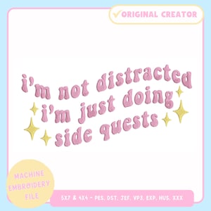 Pode incluir: Texto bordado rosa sobre fundo azul claro que diz: "Não estou distraído, estou apenas a fazer missões secundárias". Estrelas douradas acentuam o texto. A imagem é um ficheiro de bordado à máquina.