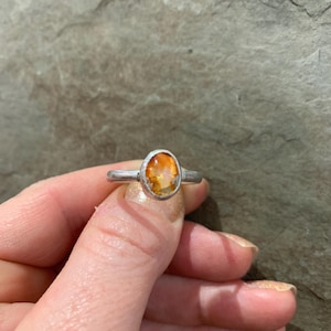 May include: A silver ring with an oval, orange gemstone. The gemstone is set in a bezel setting, and the band is a simple, thin design. The ring is held up by a hand.