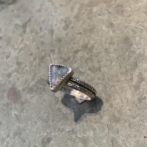 May include: A stack of silver rings. The top ring features a triangular, iridescent stone. The other rings have a textured, patterned design. The rings are displayed on a gray surface.
