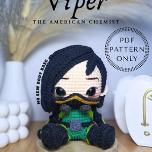 Puede incluir: Muñeca Viper de ganchillo, con un atuendo negro y verde, una máscara amarilla y cabello negro. La muñeca está sentada sobre una base de madera. El texto de la imagen dice "Viper The American Chemist" y "PDF Pattern Only".