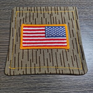 May include: A square fabric patch featuring a tan and brown vertical rain camouflage pattern. Centered on the patch is a rectangular embroidered American flag with red and white stripes and a blue field with white stars, bordered by yellow thread.