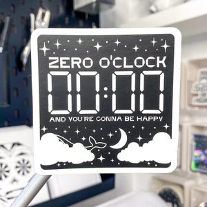 Op de afbeelding: Een zwart-witte sticker met de tekst "ZERO O'CLOCK 00:00 AND YOU'RE GONNA BE HAPPY". De sticker heeft een sterrenhemelontwerp, een maan, wolken en een walvisstaart.