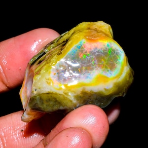 Op de afbeelding: Een ruwe, ongeslepen opaal edelsteen met een levendige kleurenpracht. De opaal heeft een mix van gele, groene, blauwe en oranje tinten, met een glinsterend, iriserend effect. De steen wordt tegen een donkere achtergrond gehouden.