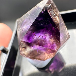 May include: A purple amethyst crystal with a clear, faceted point and a natural, rough base. The crystal has a unique, layered appearance with a hint of brown.