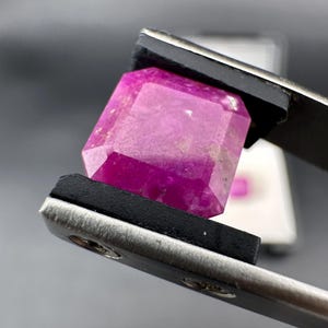 May include: A magenta-coloured, faceted gemstone is held by tweezers. The gem is rectangular with multiple facets, reflecting light. The tweezers are silver with black grips, and the background is a blurred grey.