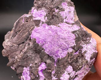 Könnte beinhalten: Ein rohes, natürliches Mineralexemplar mit einem auffälligen Farbkontrast. Der Stein zeigt eine Mischung aus tiefem Anthrazitgrau und leuchtenden Lavendeltönen. Die Lavendelbereiche haben eine faserige Textur, während die grauen Abschnitte rauer sind.